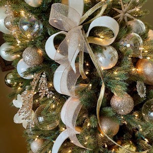 May include: A decorated Christmas tree with white, gold, and clear ornaments. A large white and gold ribbon bow is the focal point. The tree is lit with small white lights, creating a festive holiday display.