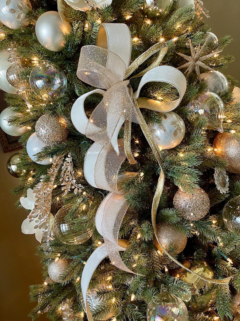 May include: A decorated Christmas tree with white, gold, and clear ornaments. A large white and gold ribbon bow is the focal point. The tree is lit with small white lights, creating a festive holiday display.