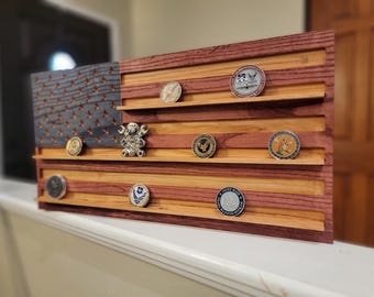 Op de afbeelding: Een houten Amerikaanse vlaggenstandaard met sleuven om challenge-munten te tonen. De vlag is gemaakt van rood, wit en blauw hout en heeft een verweerde uitstraling. Er zijn verschillende challenge-munten in de sleuven tentoongesteld.