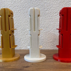 Könnte beinhalten: Drei 3D-gedruckte Ständer in Gold, Weiß und Rot. Jeder Ständer hat eine quadratische Basis und eine hohe, schmale, rechteckige Säule mit mehreren Ebenen. Die Ständer sind in einer Reihe von links nach rechts angeordnet.