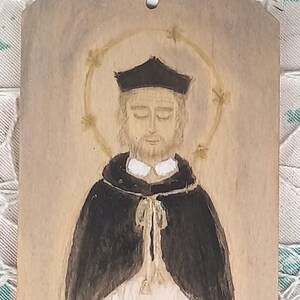 May include: A hand-painted wooden bookmark featuring a portrait of a man in a black robe and a white collar. He has a halo above his head and is depicted with his eyes closed. The background is a light wood color.