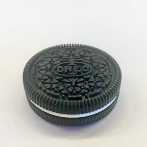 May include: A black and white Oreo cookie with the word "OREO" embossed on the top. The cookie has a textured surface and a white cream filling. The cookie is round and sits on a white surface.