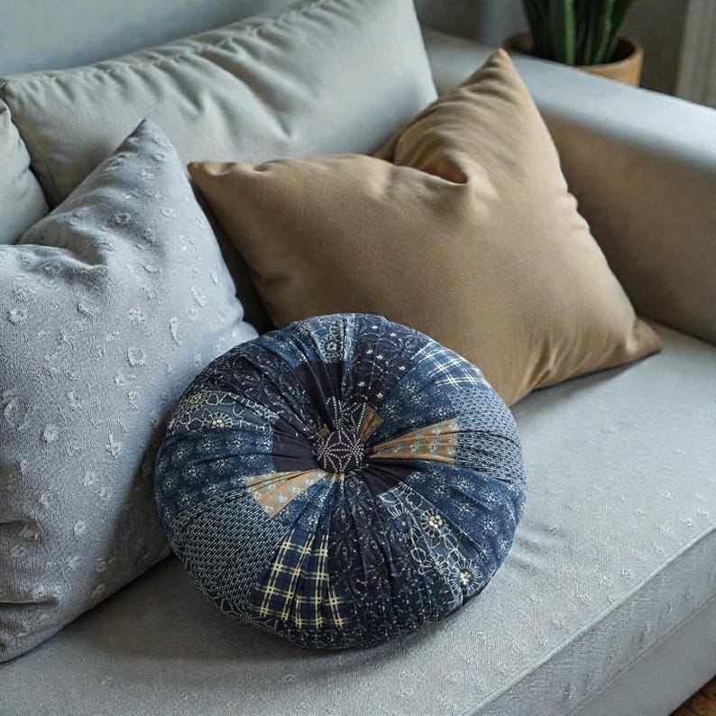 May include: A round, patchwork cushion in shades of blue, tan, and white sits on a light gray sofa, with two solid-colored pillows and a patterned pillow behind it. The cushion has a central, button-like detail.