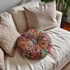May include: A round, patterned pillow with a vibrant mix of colors, including red, green, and yellow, sits on a cream-colored sofa. The pillow has a buttoned center and is surrounded by neutral-toned cushions.
