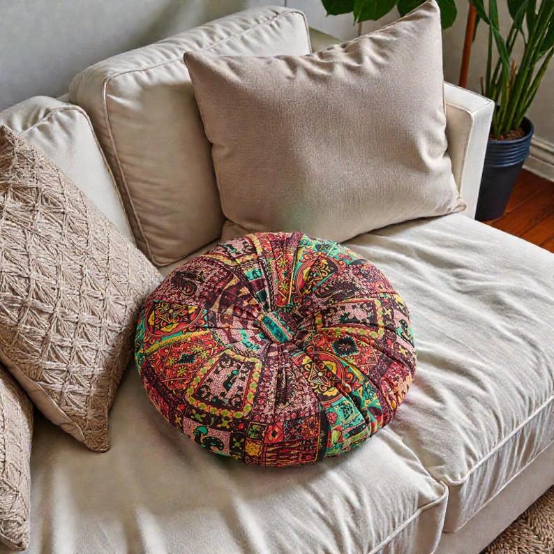 May include: A round, patterned pillow with a vibrant mix of colors, including red, green, and yellow, sits on a cream-colored sofa. The pillow has a buttoned center and is surrounded by neutral-toned cushions.