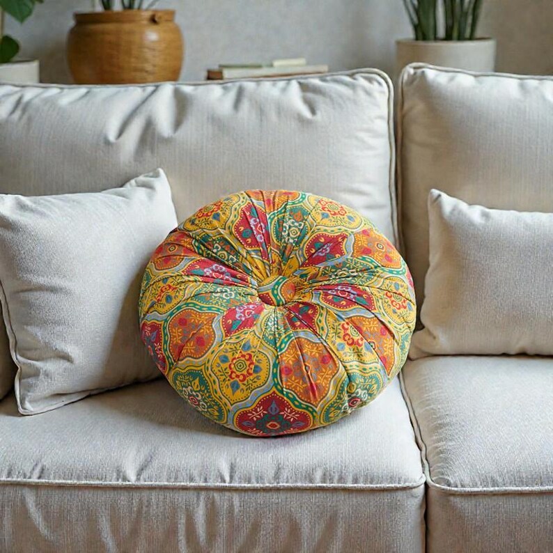 May include: A round, patterned cushion with a button center rests on a cream-colored sofa. The cushion features a vibrant design with red, yellow, green, and orange floral and geometric patterns. Two white throw pillows are on the sofa.