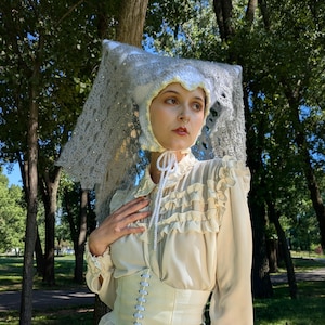 Puede incluir: Un conjunto color crema con un tocado de ganchillo con velo, una blusa con volantes y un corsé. El tocado es blanco con un delicado diseño de encaje. La blusa tiene detalles de volantes y el corsé está atado por delante.