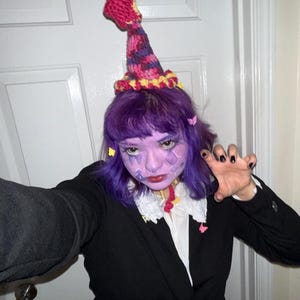 May include: A black blazer with a white collared shirt and a decorative, colorful crochet hat. The hat is primarily pink, purple, and yellow. The person in the image has purple hair and face paint.