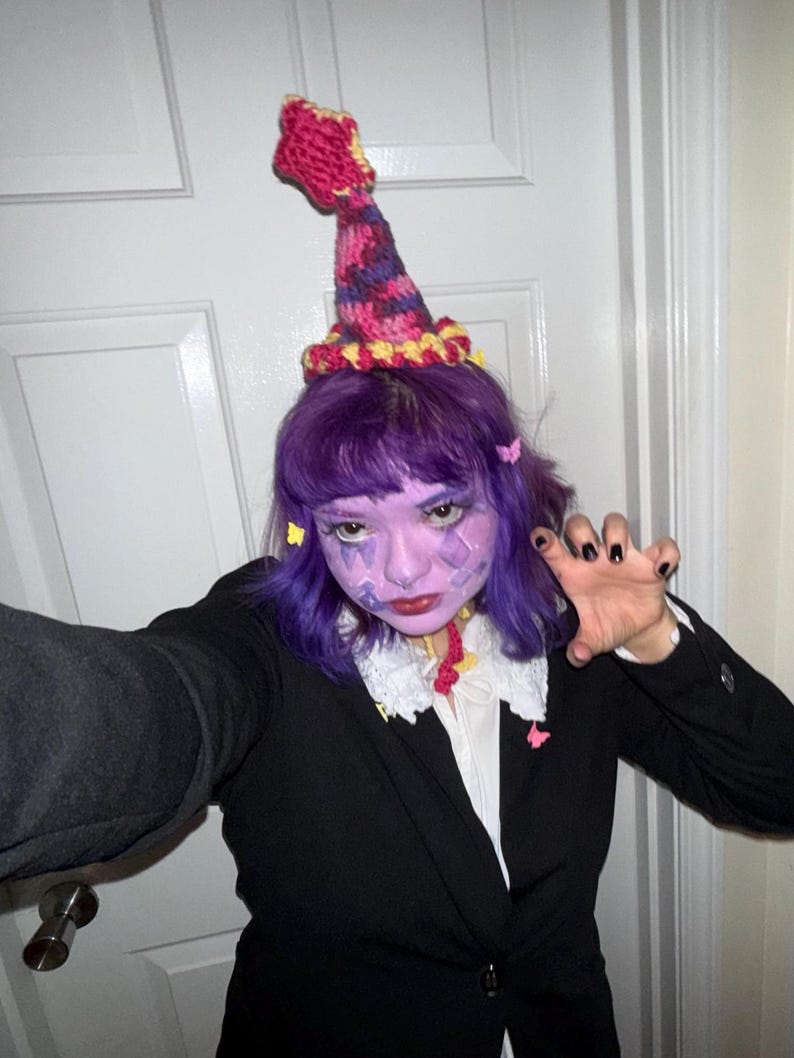 May include: A black blazer with a white collared shirt and a decorative, colorful crochet hat. The hat is primarily pink, purple, and yellow. The person in the image has purple hair and face paint.