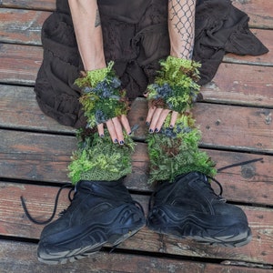 May include: A pair of green, brown, and gray fingerless gloves with a fuzzy texture. The gloves are worn by a person wearing black platform shoes.