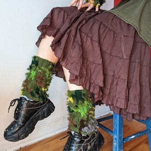 May include: A person wearing a brown skirt and black platform shoes with green and yellow camouflage socks. The socks are crocheted and have a fuzzy texture.