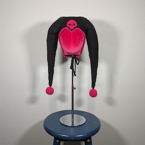 May include: A black and pink crocheted jester hat with a skull design. The hat has long, pointed ends with pink pom-poms and a pink heart-shaped centre. The hat is displayed on a stand against a neutral background.
