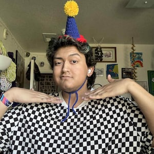 May include: A blue, red, and yellow crocheted party hat with a yellow pom-pom on top. The hat is worn with a black and white checkered shirt.