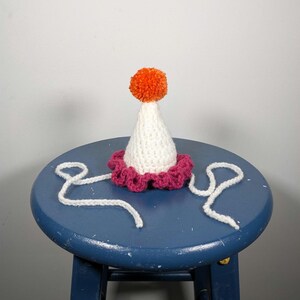 May include: A handmade party hat with a white cone, a magenta scalloped brim, and an orange pom-pom on top. White yarn ties extend from the base. The hat is sitting on a blue stool.