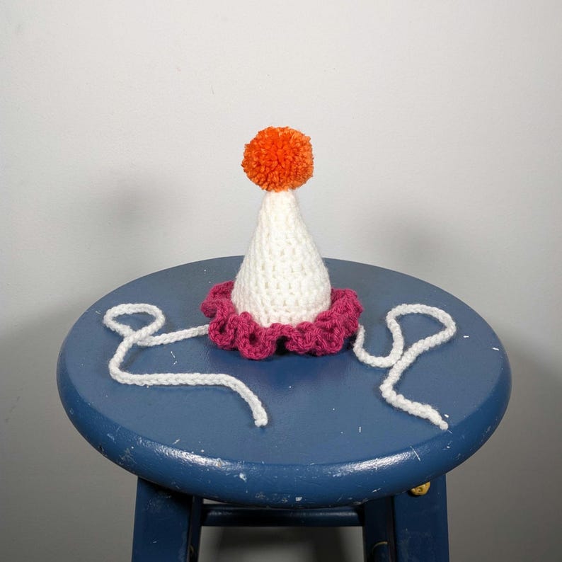 May include: A handmade party hat with a white cone, a magenta scalloped brim, and an orange pom-pom on top. White yarn ties extend from the base. The hat is sitting on a blue stool.