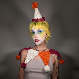 May include: A person wearing a handmade clown costume, including a white and orange crochet collar with pom-poms and a matching hat. The hat is white with a pink trim and an orange pom-pom on top. The person has yellow hair and clown makeup.