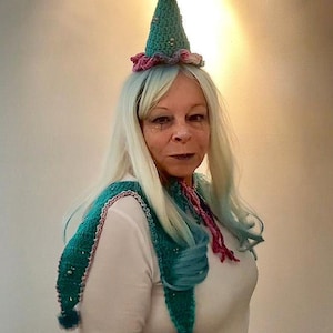 May include: A teal and pink crocheted jester hat with a matching scarf. The hat has a maroon pom-pom on top and is adorned with small gold beads. The scarf has teal and pink accents and pom-poms at the ends.