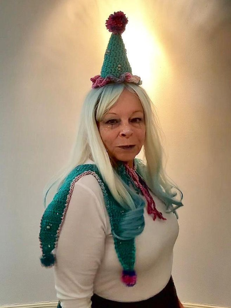 May include: A teal and pink crocheted jester hat with a matching scarf. The hat has a maroon pom-pom on top and is adorned with small gold beads. The scarf has teal and pink accents and pom-poms at the ends.