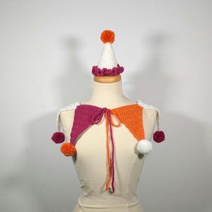 May include: A handmade crochet jester collar and hat set. The collar features magenta and orange triangles with white accents and pom-poms. The hat is white with an orange pom-pom and a magenta trim. The set is displayed on a mannequin.