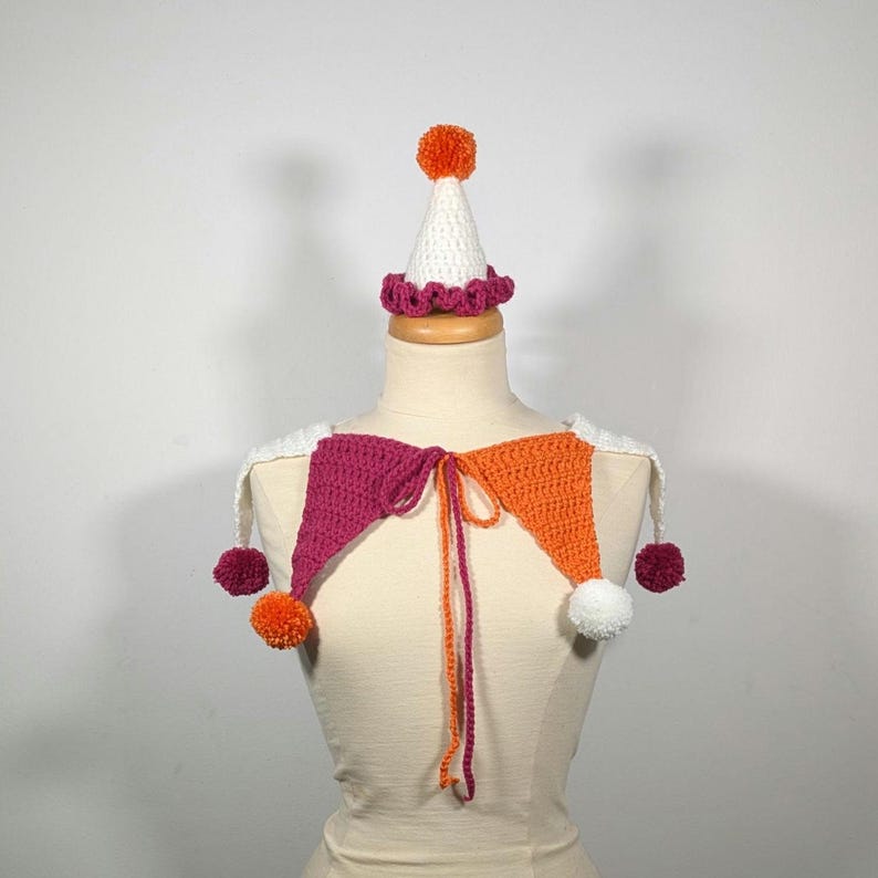 May include: A handmade crochet jester collar and hat set. The collar features magenta and orange triangles with white accents and pom-poms. The hat is white with an orange pom-pom and a magenta trim. The set is displayed on a mannequin.