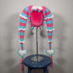 May include: A handmade crochet hat in pink and turquoise stripes with white trim and heart-shaped accents. The hat has long, pointed extensions with white ruffles and dangling heart-shaped ornaments. The hat is displayed on a pink mannequin head.