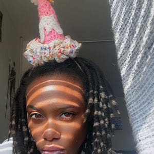 May include: A handmade party hat with a pink cone, white frosting-like accents, and a colorful pom-pom. The base of the hat is a ruffled design with flecks of color. The hat is designed to be worn on the head.