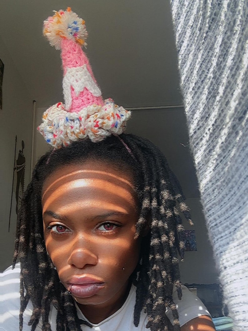 May include: A handmade party hat with a pink cone, white frosting-like accents, and a colorful pom-pom. The base of the hat is a ruffled design with flecks of color. The hat is designed to be worn on the head.
