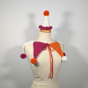 May include: A handmade crochet jester collar and hat set. The collar features magenta and orange diamond shapes with white accents and pom-poms. The hat is white with a magenta ruffle and orange pom-pom. The set is displayed on a mannequin.
