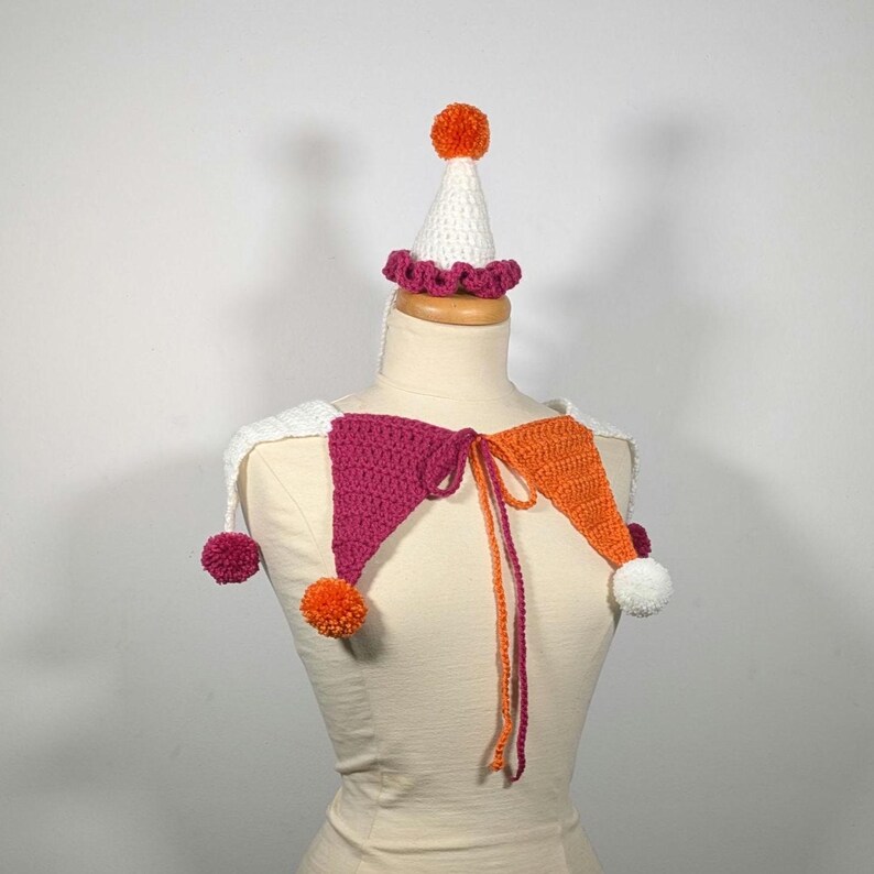 May include: A handmade crochet jester collar and hat set. The collar features magenta and orange diamond shapes with white accents and pom-poms. The hat is white with a magenta ruffle and orange pom-pom. The set is displayed on a mannequin.