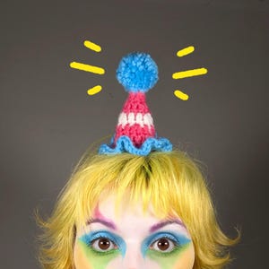 May include: A handmade party hat in pink, white, and blue, with a blue pom-pom and ruffled brim. The hat sits on yellow hair, with a face below featuring bright makeup. Yellow starbursts are drawn around the hat.
