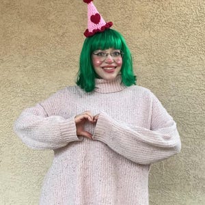 May include: A pink and red crochet party hat with a heart design and a red pom-pom. The hat is worn with a green wig, glasses, and a pink sweater. The person is making a heart shape with their hands.
