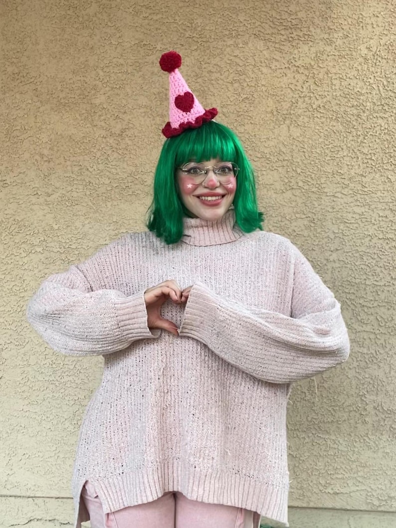May include: A pink and red crochet party hat with a heart design and a red pom-pom. The hat is worn with a green wig, glasses, and a pink sweater. The person is making a heart shape with their hands.