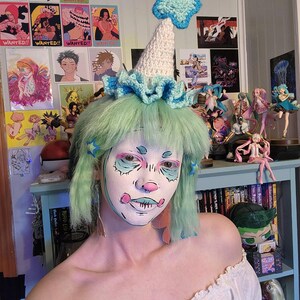 May include: A whimsical, handmade crochet hat with a light blue star and trim. The hat is white and cone-shaped, with a ruffled brim. The person in the image has green hair and face paint, and is wearing an off-the-shoulder white top.