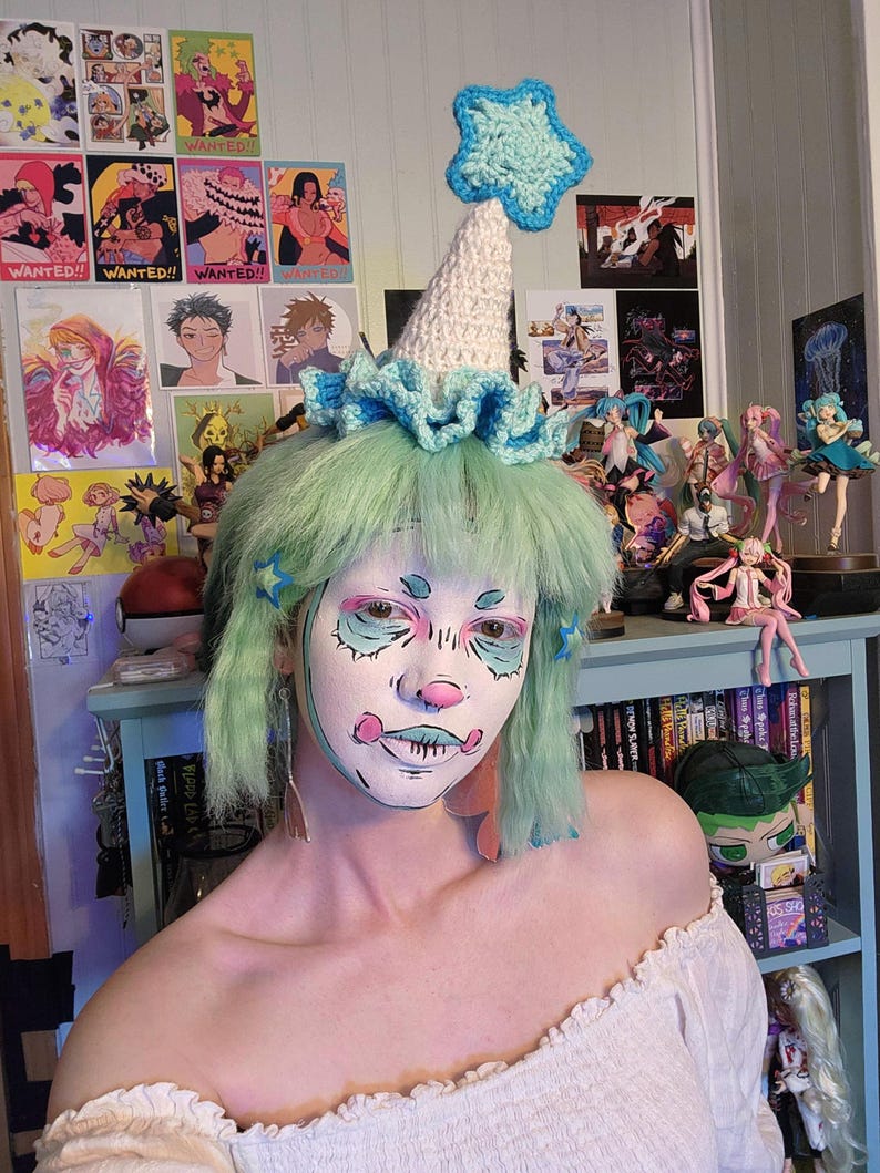 May include: A whimsical, handmade crochet hat with a light blue star and trim. The hat is white and cone-shaped, with a ruffled brim. The person in the image has green hair and face paint, and is wearing an off-the-shoulder white top.