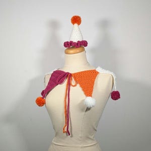 May include: A handmade crochet jester collar and hat set. The collar features magenta, orange, and white triangles with matching pom-poms. The hat is white with a magenta trim and an orange pom-pom on top. The set is displayed on a mannequin.