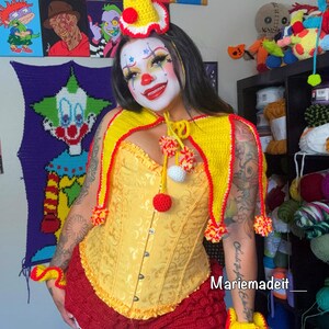 May include: A yellow and red crochet clown costume. The costume includes a yellow corset, a red ruffled skirt, a yellow cape with red trim, and a matching hat. The image also includes yellow and red wrist cuffs. The text "Mariemadeit_" is visible.
