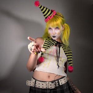 May include: A whimsical outfit featuring a black and yellow striped hat with a pink trim and pom-pom. The outfit includes a matching scarf and a white crop top. The person is wearing a plaid skirt and a white belt.