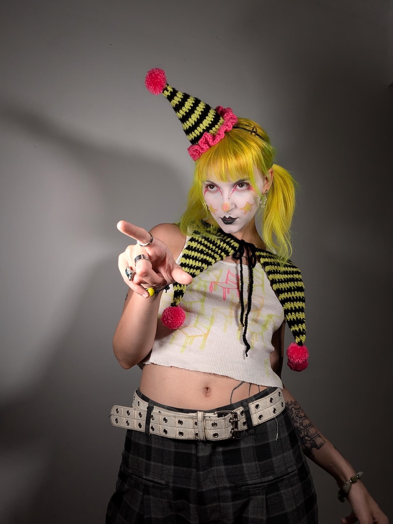 May include: A whimsical outfit featuring a black and yellow striped hat with a pink trim and pom-pom. The outfit includes a matching scarf and a white crop top. The person is wearing a plaid skirt and a white belt.