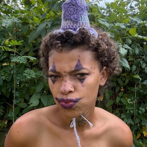 May include: A handmade crochet hat in shades of purple and blue, with a star-shaped design. The hat has a ruffled brim and a pointed top. The person in the image has face paint and a white string around their neck.