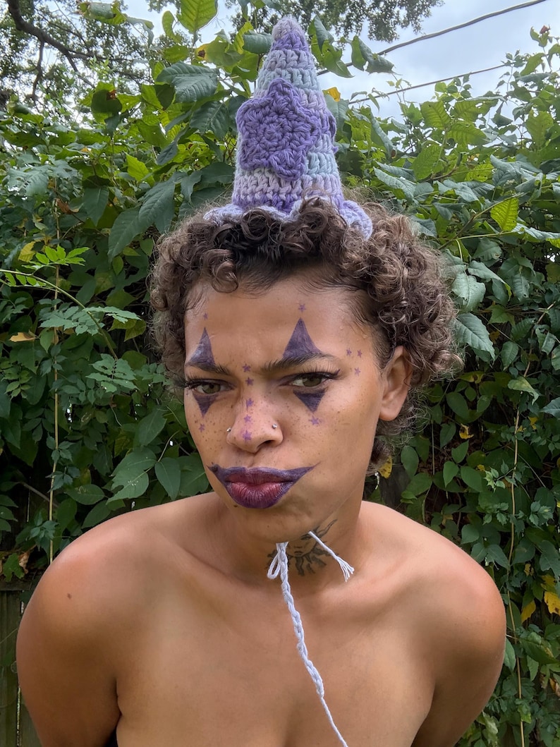 May include: A handmade crochet hat in shades of purple and blue, with a star-shaped design. The hat has a ruffled brim and a pointed top. The person in the image has face paint and a white string around their neck.