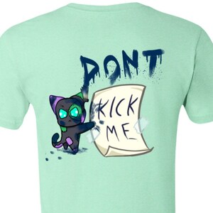 Pode incluir: Uma camiseta verde claro com um gato de desenho animado que está pintando com spray as palavras "DON'T KICK ME" em um pedaço de papel.