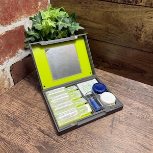 May include: A gray contact lens travel case with a lime green interior and a built-in square mirror. The kit includes four small clear plastic vials of solution, a blue and clear lens case, and a white screw-top container for lens storage.