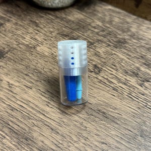 May include: A small, clear plastic container with a white, ridged cap and blue accents. Inside are two blue objects, one a darker shade than the other. The container is on a wooden surface.