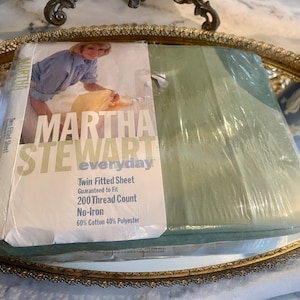 May include: A new, packaged twin fitted sheet by Martha Stewart Everyday. The package is clear, showing a light green sheet inside. The label features a photo of a woman and text that reads "Twin Fitted Sheet, 200 Thread Count, No-Iron, 60% Cotton 40% Polyester."