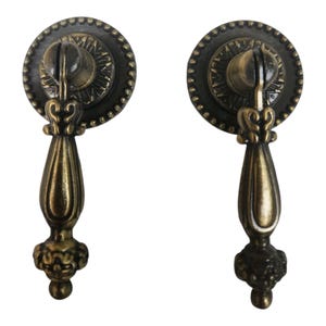 Op de afbeelding: Twee antieke bronzen lade-knoppen. Elke knop heeft een cirkelvormige achterplaat met een parelrand en een centraal ontwerp. De druppelvormige handgreep is opgehangen aan de achterplaat, met sierlijke details.