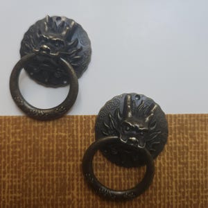 以下が含まれることがあります： 2つのアンティークブロンズ色のキャビネットプル。各プルは、詳細なドラゴンのデザインが施された丸いバックプレートに取り付けられた円形のリングハンドルを備えています。プルは、白とテクスチャード加工された茶色の背景に設定されています。