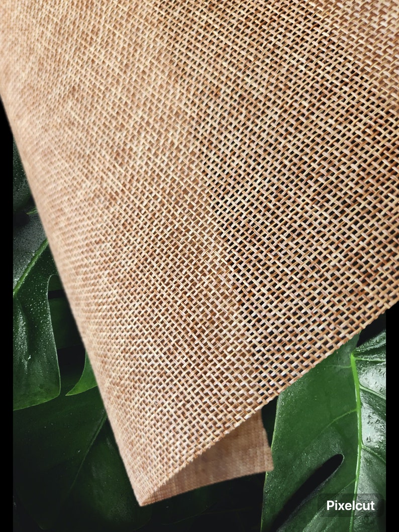 May include: Close-up of a beige burlap fabric with a woven texture. The fabric is folded and partially obscured by a green leaf.
