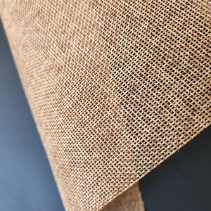 May include: Close-up of a natural brown burlap fabric with a woven texture. The fabric is folded over on itself, showing the underside.