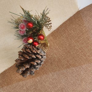 May include: A brown pine cone and a sprig of greenery with red berries are arranged on a beige and brown burlap fabric.