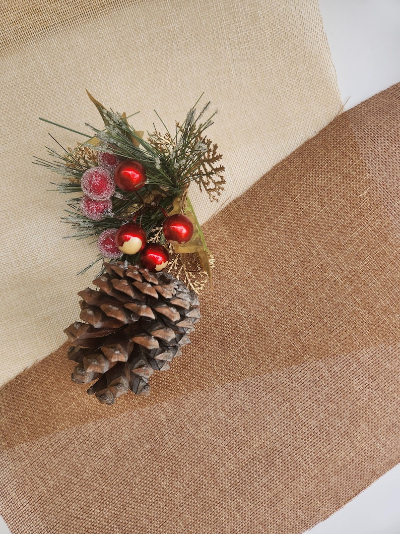 May include: A brown pine cone and a sprig of greenery with red berries are arranged on a beige and brown burlap fabric.